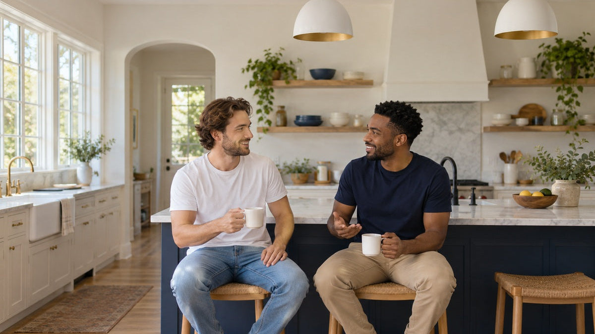Illustrative unbranded scene of two men in plain white and navy T-shirts talking in a bright home kitchen.