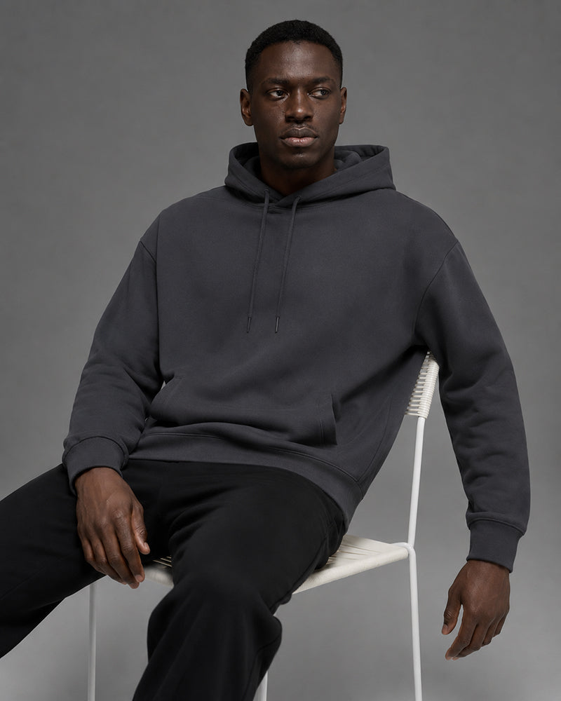Man in a charcoal hoodie walking through light rain on a city street, hood up, hands in pockets, fall leaves on the sidewalk
