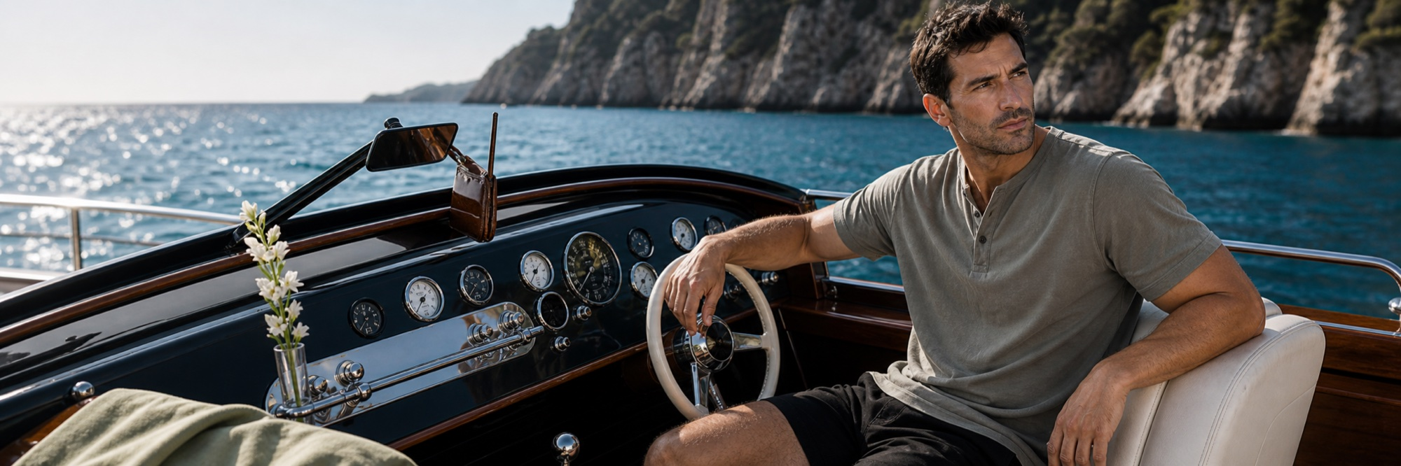 Man at the helm of a vintage boat, olive Henley tee, sunny coastal water